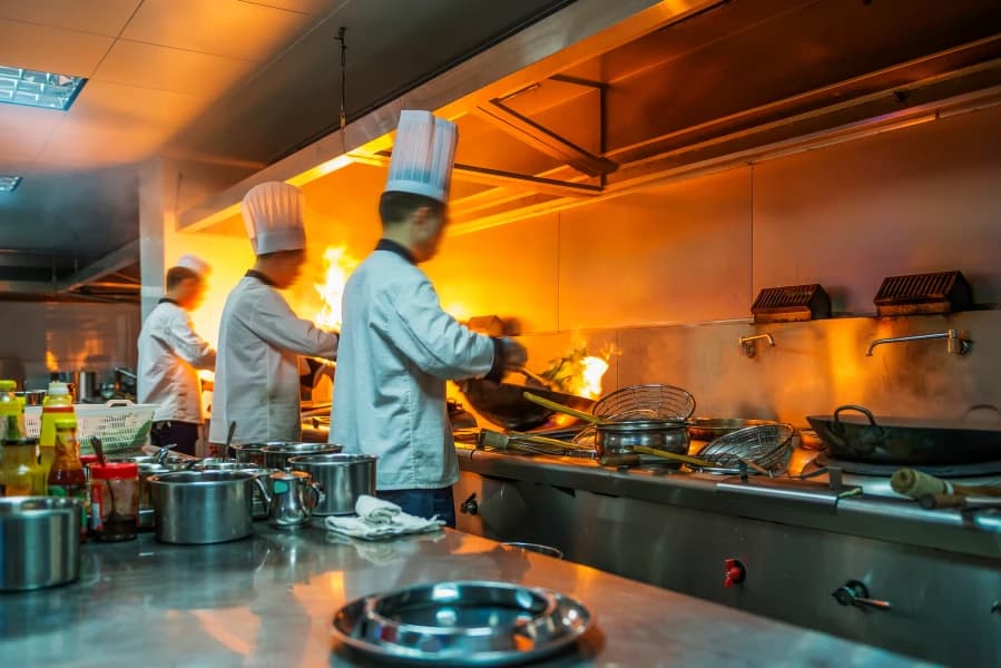 Chefs cooking in a busy commercial kitchen environment with open flames and professional kitchen equipment in use.