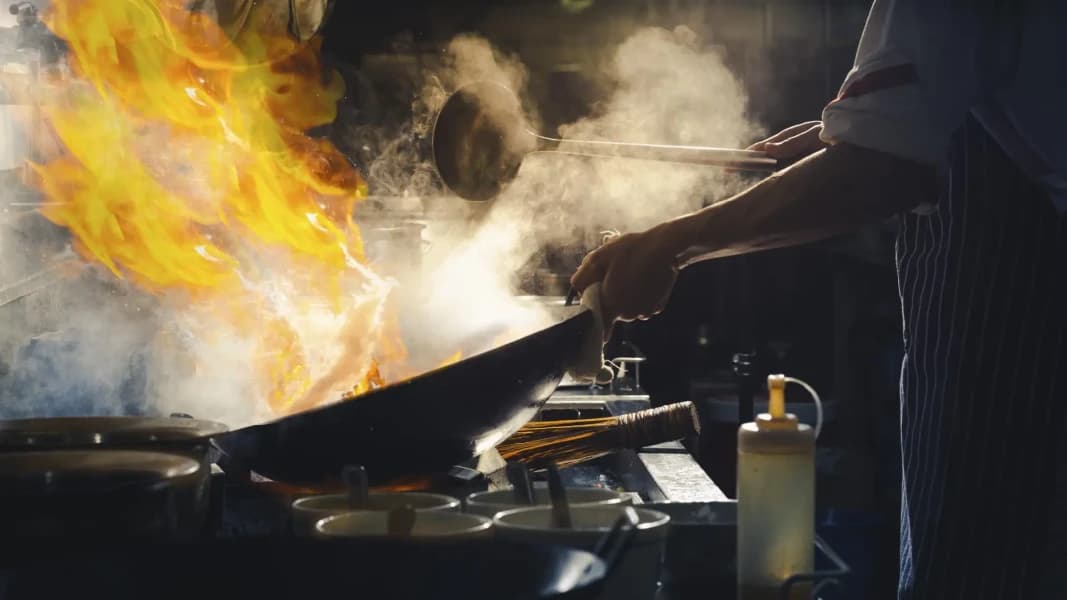 Chef cooking in a commercial kitchen with wok and visible smoke, showcasing busy hospitality environment.